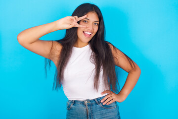 Fototapeta na wymiar Young hispanic girl wearing tank top over blue background making v-sign near eyes. Leisure, coquettish, celebration, and flirt concept.