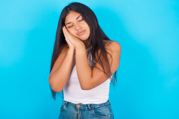 Fototapeta na wymiar Relax and sleep time. Tired Young hispanic girl wearing tank top over blue background with closed eyes leaning on palms making sleeping gesture.