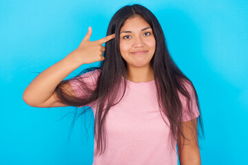 Unhappy Young hispanic girl wearing pink T-shirt over blue background curves lips and makes suicide...