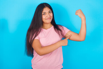 Obraz na płótnie Canvas Smiling Young hispanic girl wearing pink T-shirt over blue background raises hand to show muscles, feels confident in victory, strong and independent.