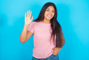 Young hispanic girl wearing pink T-shirt over blue background waiving saying hello or goodbye happy and smiling, friendly welcome gesture.