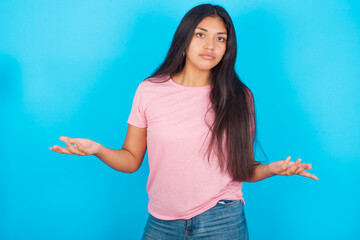 Young hispanic girl wearing pink T-shirt over blue background  looks uncertain shrugs shoulders.