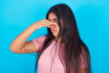 Displeased Young hispanic girl wearing pink T-shirt over blue background plugs nose as smells...