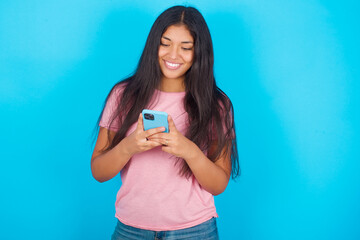 Smiling Young hispanic girl wearing pink T-shirt over blue background using cell phone, messaging,...