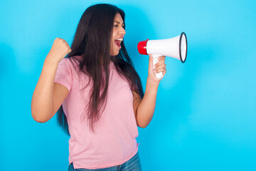 Young hispanic girl wearing pink T-shirt over blue background communicates shouting loud holding a megaphone, expressing success and positive concept, idea for marketing or sales.