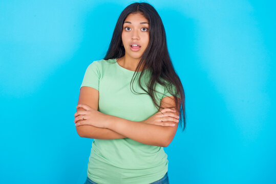 Shocked embarrassed Young hispanic girl wearing green T-shirt over blue background keeps mouth widely opened. Hears unbelievable novelty stares in stupor