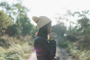 Happy beautiful young adventurous woman in casual clothes with camera and wool bobble hat, warms her hand by breathing hand to make warm in forest while walking and hiking for camping on epic mountain