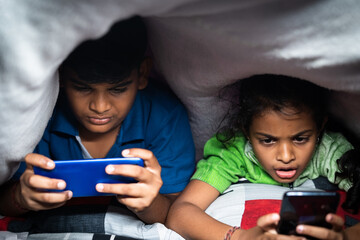 Kids busy playing video game on mobile phone at night under the bedsheet - concept smartphone and...