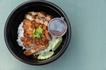 Closeup of food as Crispy fried pork belly with Thai spicy sauce, cucumber on white jasmine rice in black modern bowl on clean green minimal clolor  table cloth background and texture with copy space.