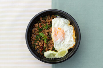 Thai traditional food, Flavorful appetizing food as Spicy fried pork with basil leaves, fried egg and cucumber in black modern bowl on clean minimal cream and green color cloth background. Copy space.