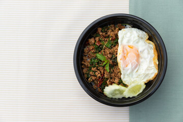 Thai traditional food, Flavorful appetizing food as Spicy fried pork with basil leaves, fried egg and cucumber in black modern bowl on clean minimal cream and green color cloth background. Copy space.