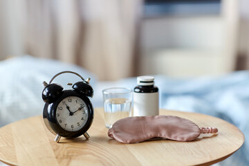 sleep disorder, bedtime and morning concept - close up of alarm clock, eye sleeping mask, glass of water and soporific medicine on night table at home
