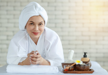Asian happy relax female model in white clean bathrobe lay down on massage bed with compress ball coconut oil and plumeria flower in tray smiling using smartphone waiting for body massaging therapy