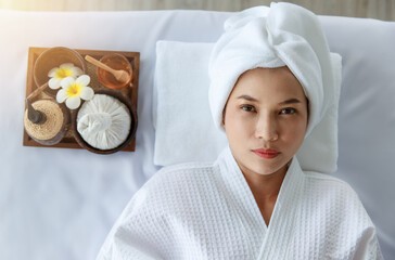 Close up facial shot of Asian happy female model in white clean bathrobe lying down closed eyes relaxing on massage bed with compress ball coconut oil and plumeria in tray smiling wait for therapy