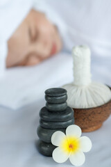 Fototapeta na wymiar Close up shot white yellow plumeria flower bloom with oil and hot stones compress stuff in towel placing on massage bed while spa therapy Asian female client lay down relaxing in blurred background