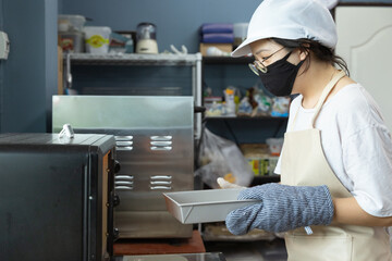 Happy young Asian woman with protective face mask, apron is smiling while bringing bakery material to oven to bake homemade cake or bread in home kitchen, learning new skills when home quarantine.