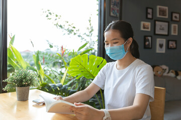 Happy young Asian woman with medical protective face mask, white casual t-shirt is smiling, reading book in cafe, coffee shop with little cute green tree with social distancing, new normal lifestyle.