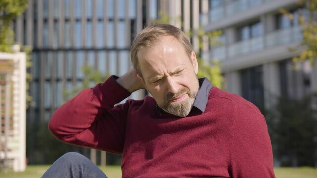 A middle-aged handsome Caucasian man thinks about something as he sits in a street in an urban area - office buildings in the blurry background - closeup