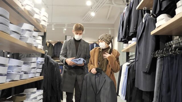 Beautiful young couple in medical masks holding a jacket and golf go to the fitting room in the mall of men's clothing boutique. Man and woman go shopping during seasonal discounts. Sale time concept.