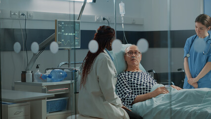 Nurse and african american doctor tending to ill patient in hospital ward bed at medical facility. Sick old man with nasal oxygen tube and IV drip bag recovering from injury and disease