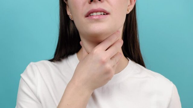 Close up of sick young woman suffering from sore throat, isolated on blue studio wall. Causes of throat pain include flu, common cold, bacterial infections, pneumonia or allergies. Medical concept
