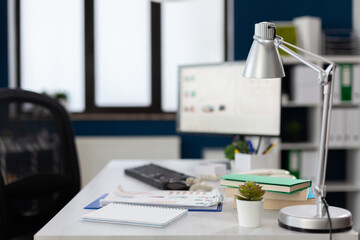 Closeup of lamp on white desk in empty modern business office. Notebook and clipboard with papers on table in startup workspace. Computer screen in indoors modern commercial workplace.
