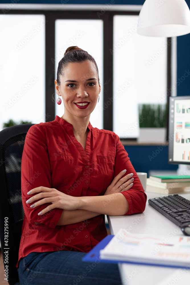 Wall mural Portrait of entrepreneur sitting at desk in startup office. Businesswoman in red shirt looking confident in business workspace. Successful employee posing with crossed arms in front of desktop