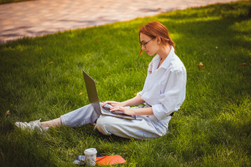 beautiful girl studies and works with laptop, blogger, distance education while sitting on the grass