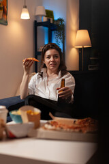 Happy person after work looking at television sitcom show eating a slice of hot pizza delivery sitting on couch holding beer bottle. Woman enjoying takeaway tv dinner on sofa.