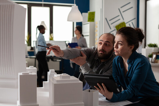 Architect Pointing Pen At Skyscraper Architectural Model Collaborating With Colleague Engineer Holding Tablet With Blueprints In Modern Office. Architects Looking Inspired Collaborating On Maquette.