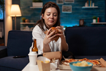Woman sitting on couch looking at tasty delicious burger delivery dinner in living room in front of television. Person standing at table with takeaway fast food meal large pizza.