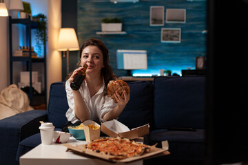 Caucasian woman drinking craft bottled beer eating delicous takeaway hamburger sitting on couch in living room. Person laughing watching television having takeout order pizza and junk food.