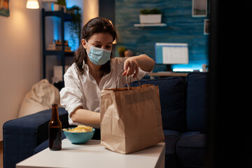 Fototapeta na wymiar Caucasian female back from office with face mask preparing to unpack takeaway bag with fast food. Woman home from work wearing covid protection preparing for delivery dinner.