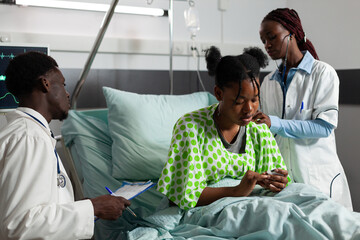 Sick patient standing in bed while african american physician doctor monitoring heart rate using medical stethoscope during clinical appointment in hospital ward, Therapist man writing treatment