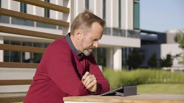 A middle-aged handsome Caucasian man looks at a tablet and thinks as he sits at a table in a park in an urban area - office buildings in the blurry background - closeup