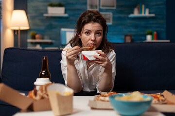 Woman eating takeout asian noodles with chopsticks on living room sofa while watching news on television. Office worker sitting on couch in the evening enjoying chinese takeaway ramen box.