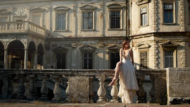 Girl posing on the background of a beautiful ancient castle sitting on the parapet. The wedding bride during a photo session. Glamorous elegant girl with beautiful sexy legs.