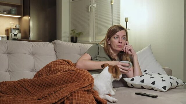 Woman with her dog lying on sofa and watching TV, drinks tea from cup. Resting at home with pet