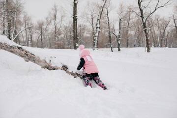 Distant photo of female Russian kid climbing tree log with efforts wearing pink winter clothes in forest. Astonishing background full of white color and snow. 