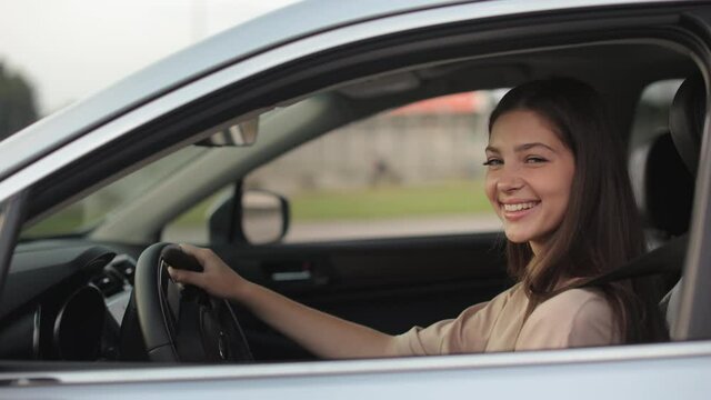 A young woman is sitting in the car and smiling. She is turning her head and looking at the camera. She is keeping her hands on the steering wheel. The camera zooms in. 4K