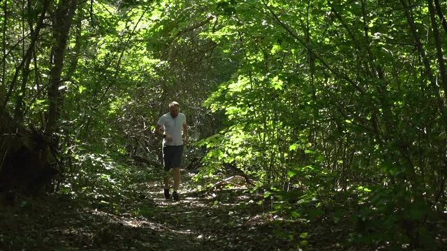 a young man in a white meek and gray shorts with headphones runs through the forest on a summer morning. the bright sun shines, hard shadows.