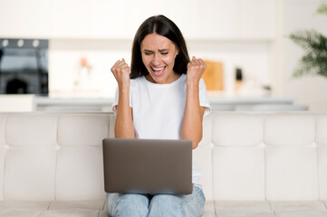 Happy young cute confident brunette caucasian woman, in casual clothes, sitting on sofa, using laptop, celebrating good profit, rejoice news or message, gesturing with fists, looking at screen, smile