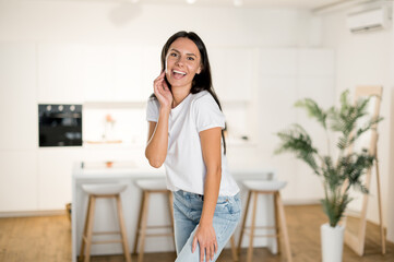 Beautiful happy cheerful Caucasian young adult brunette woman, in casual stylish clothes, stands in modern living room at home, looking at camera, smiling friendly, looks happy