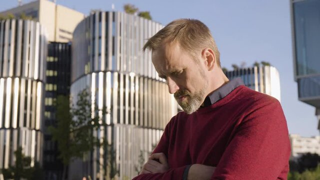 A middle-aged handsome Caucasian man thinks about something and acts sad in an urban area - office buildings in the blurry background - closeup