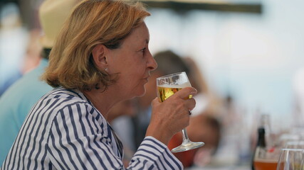 Senior woman drinking glass beer seated at restaurant. Older lady takes a sip of refreshing beverage
