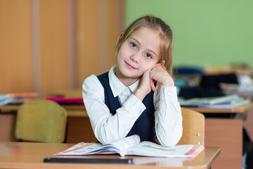A cute girl schoolgirl sits at a desk surrounded by books. Looks into the camera. School life