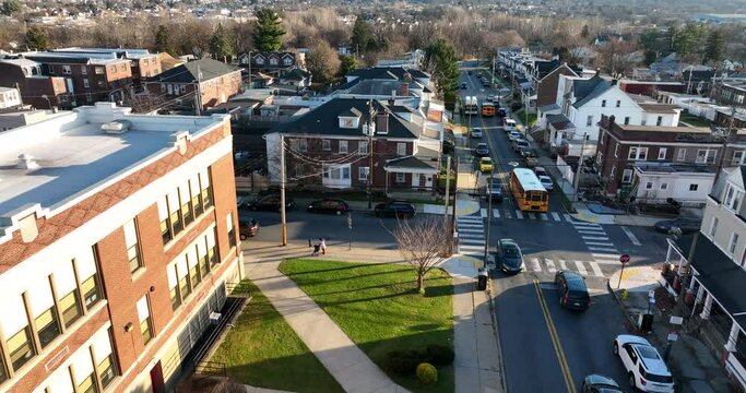 Aerial tracking shot of yellow school bus past public school building. Students live in urban city homes and houses. Bus stop in town.