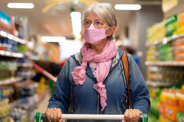 Senior woman shopping at supermarket pushing cart, wearing new ffp2 face mask more protective...