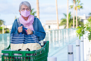 Elderly woman after shopping at the supermarket removes the bags from the trolley wearing the new most protective ffp2 mask against the omicron variant of covid-19