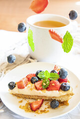 piece of strawberry and blueberry cheescake on plate and cup of tea served for breakfast at home, hotel or cafe. Berries levitating and falling down. Dessert meal, sweet food . High quality photo.